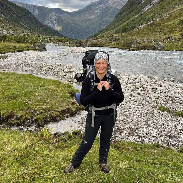 Person med tursekk står ved elv i fjellandskap omgitt av høye fjell og natur.