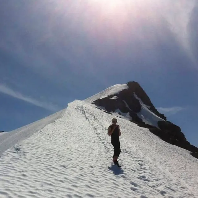 Person står på snødekt fjellrygg med utsikt mot fjelltopp under klar sommerhimmel.